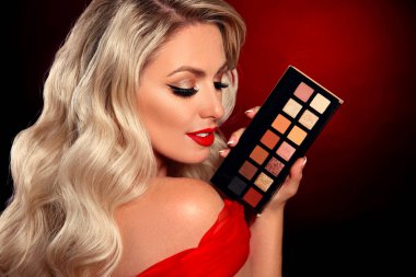 Güzellik makyajı. Kırmızı dudaklar, kozmetik göz farı paleti. Uzun dalgalı saç stili koyu arka planda ruj tutan seksi sarışın kadının moda portresi. Güzel kadın yüzü. 