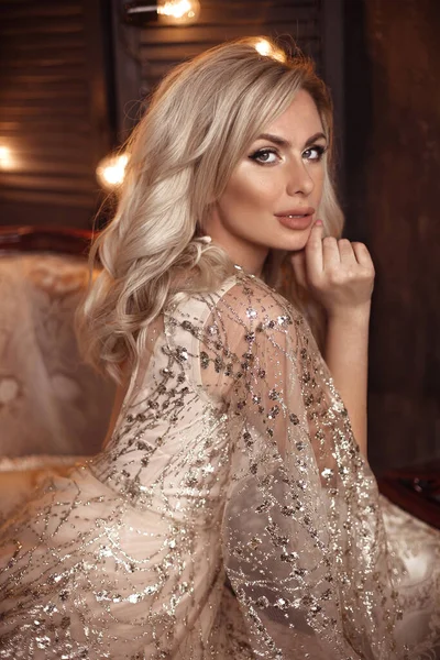 Elegant blond woman in beige dress posing on luxury sofa in royal interior. Fashion beautiful sensual bride with makeup, curly hair style in luxurious prom gown.