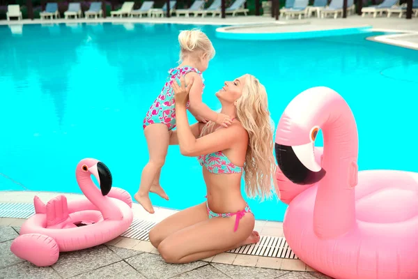 Aile görünüşü. Güzel anne kızıyla eğleniyor. İyi tatiller. Küçük kızıyla sarışın, güzel bir kadın. Pembe şişme havuz flamingosu eşliğinde aynı mayoları giyiyor..