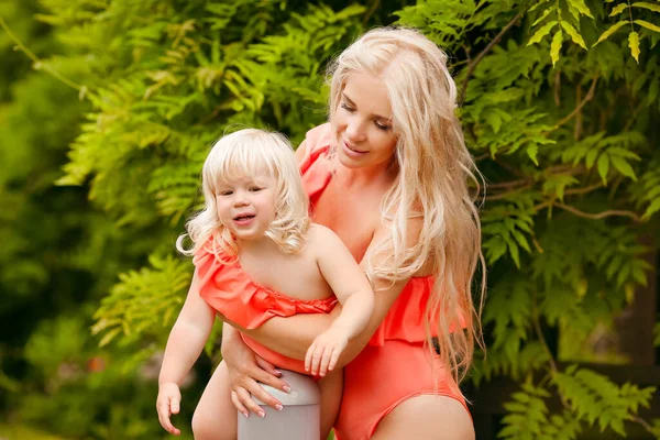家族みたいに 美しい母は緑の公園の上に彼女の小さなブロンドの娘を持っています 夏休みに かわいい女の子は楽しみを持っているファッションオレンジ水着で身に着けています 親の屋外肖像画 — ストック写真