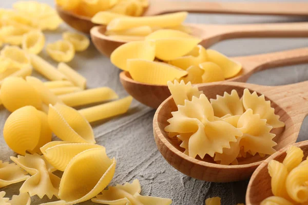 Different uncooked pasta and wooden spoons on grey table