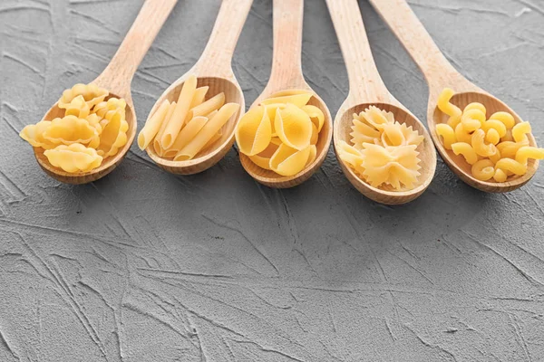 Деревянные ложки с различными сырыми макаронами на сером столе — стоковое фото