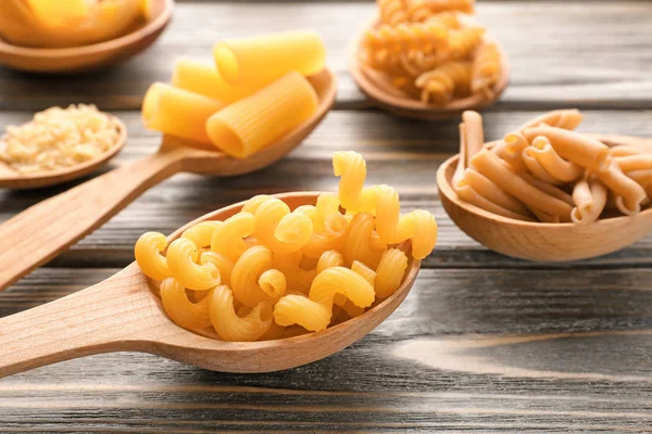 Spoons with different uncooked pasta on wooden background — Stock Photo, Image