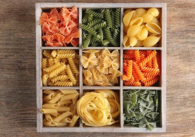 Wooden box with different raw pasta on table clipart