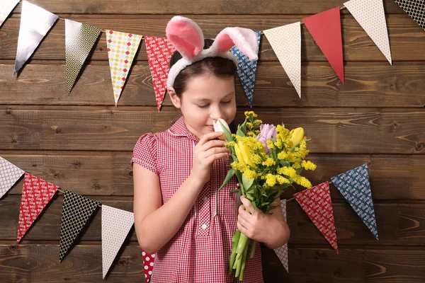 Niedliches kleines Mädchen mit Osterhasenohren und schönen Blumen in der Nähe der dekorierten Holzwand — Stockfoto