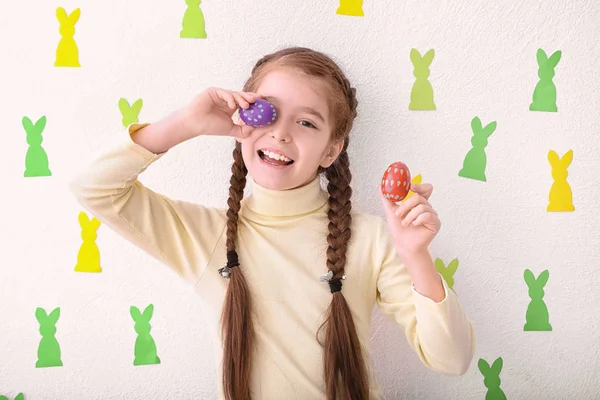 Cute little girl holding Easter eggs near white wall decorated with paper bunnies — Stock Photo, Image