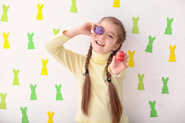 Cute little girl holding Easter eggs near white wall decorated with paper bunnies — Stock Photo, Image
