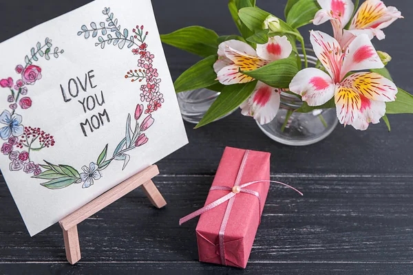 Card with phrase "Love you mom", flowers and gift box on table. Greeting for Mother's day — Stock Photo, Image