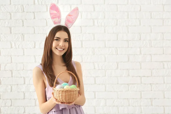 Bella giovane donna con uova di Pasqua e orecchie di coniglio contro muro di mattoni bianchi — Foto Stock