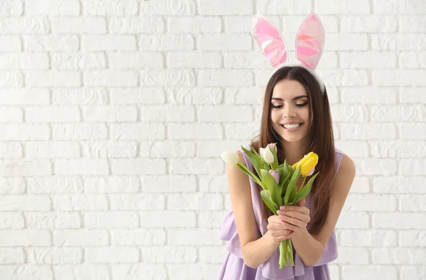 Bella giovane donna con orecchie coniglietto di Pasqua e mazzo di fiori contro muro di mattoni bianchi — Foto Stock