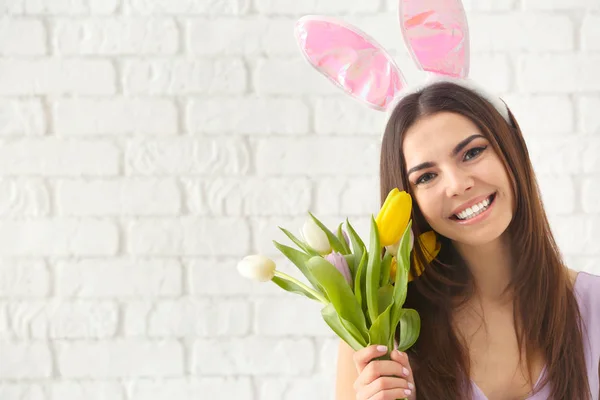 Bella giovane donna con orecchie coniglietto di Pasqua e mazzo di fiori contro muro di mattoni bianchi — Foto Stock