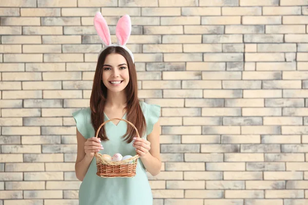 Beautiful young woman with Easter eggs and bunny ears against brick wall — Stock Photo, Image