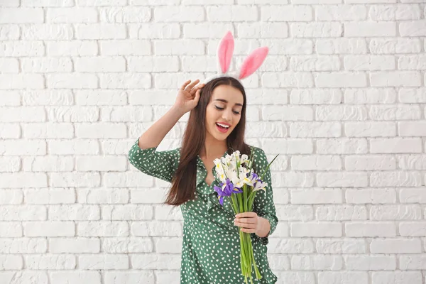 Vacker ung kvinna med Easter bunny öron och bukett blommor mot vit tegelvägg — Stockfoto