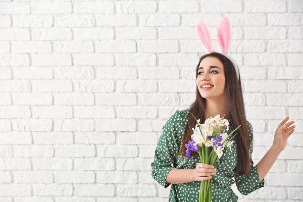 Mooie jonge vrouw met Easter bunny oren en boeket bloemen tegen witte bakstenen muur — Stockfoto