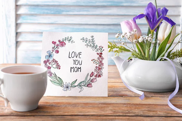 Card with words LOVE YOU MOM, cup of tea and flowers on table. Mother's day celebration — Stock Photo, Image