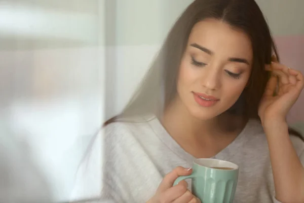 美丽的年轻女子与杯热茶在家里, 透过窗口查看 — 图库照片