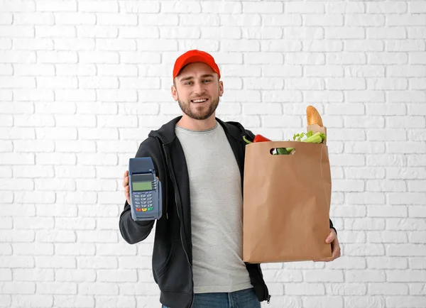 Молодой человек с бумажным пакетом и банковским терминалом на фоне кирпичной стены. Доставка продуктов питания — стоковое фото