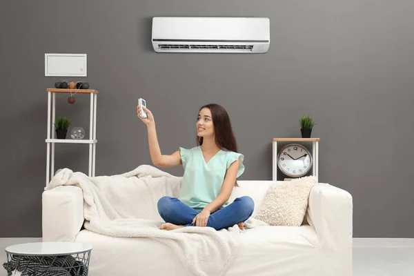 Young woman switching on air conditioner while sitting on sofa at home