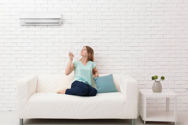 Young woman switching on air conditioner while sitting on sofa at home