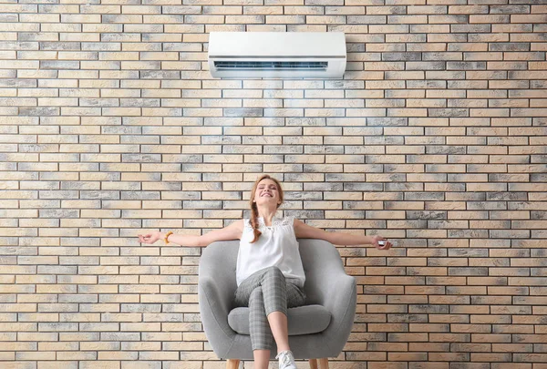 Mujer joven sentada en sillón y disfrutando del flujo de aire fresco desde el acondicionador en el interior — Foto de Stock