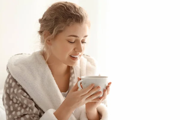 Young woman in robe drinking tea on light background — Stock Photo, Image
