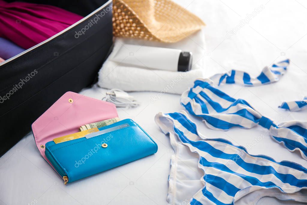 Wallet, swimsuit and open suitcase with packed things on bed