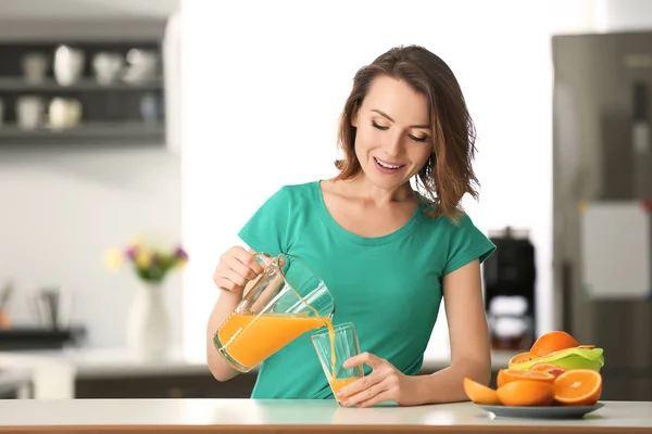 Beautiful woman pouring citrus juice from jug into glass in kitchen