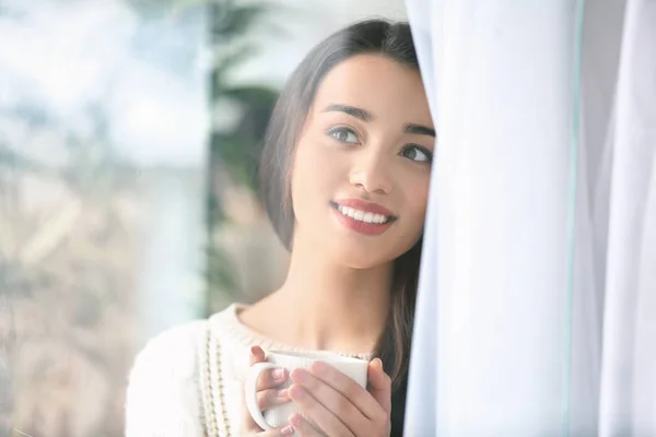 Beautiful young woman with cup of hot tea at home, view through window — Stock Photo, Image
