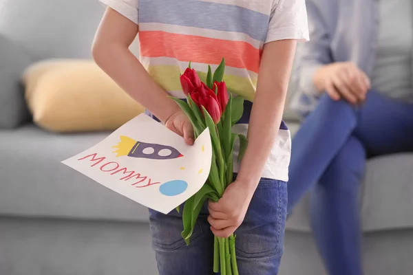 Little boy hiding gifts for mother behind his back at home — Stock Photo, Image