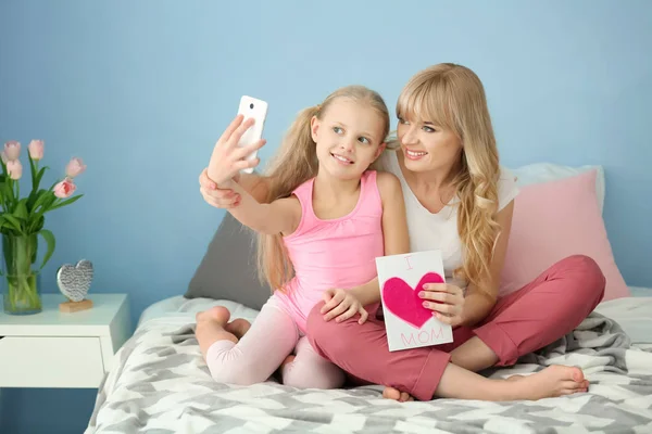 Cute little girl and her mother taking selfie with handmade card in bedroom — Stock Photo, Image