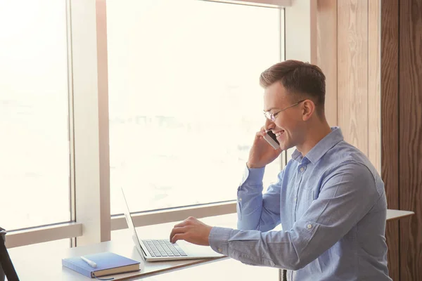 Young freelancer talking on mobile phone while working with laptop in cafe — Stock Photo, Image