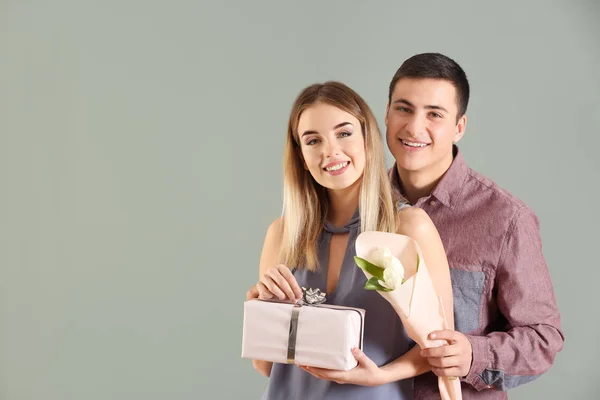 Jovem casal feliz com presente e flores no fundo de cor — Fotografia de Stock