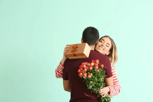 Happy young woman with gift box and flowers embracing her boyfriend on color background — Stock Photo, Image