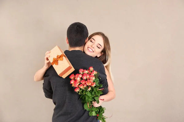 Šťastná mladá žena s dárkovou krabičku a květiny objímá svého přítele na barvu pozadí — Stock fotografie