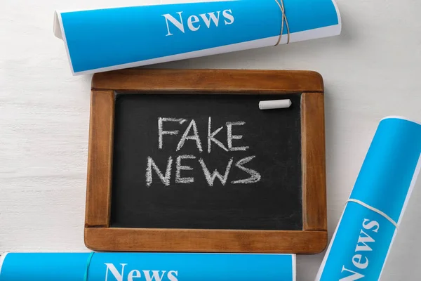 Chalkboard with text FAKE NEWS and paper rolls on wooden background — Stock Photo, Image