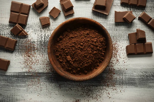 Bowl with cocoa powder and tasty chocolate on wooden background, top view