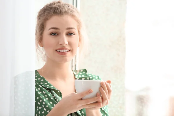 Młoda kobieta picia herbaty w pobliżu okna w pomieszczeniu — Zdjęcie stockowe