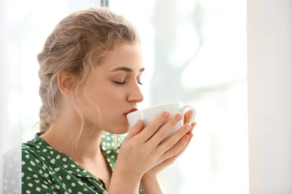 Mujer joven bebiendo té cerca de la ventana interior — Foto de Stock