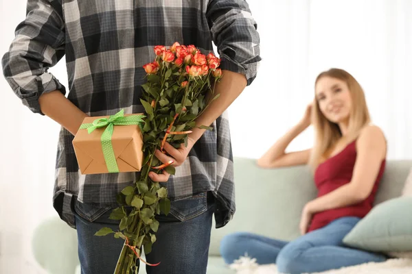 Young man hiding gift and flowers for girlfriend behind his back at home — Stock Photo, Image