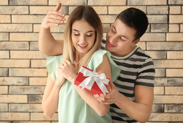 Young man giving present to his beloved girlfriend near brick wall — Stock Photo, Image
