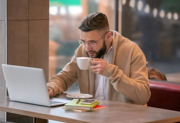 Молодий фрілансер з чашкою кави і ноутбуком, що працює в кафе — стокове фото
