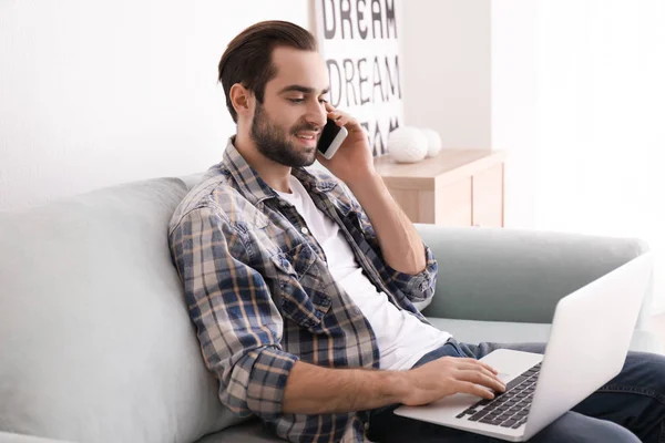 Male freelancer talking on phone while working with laptop in home office — Stock Photo, Image