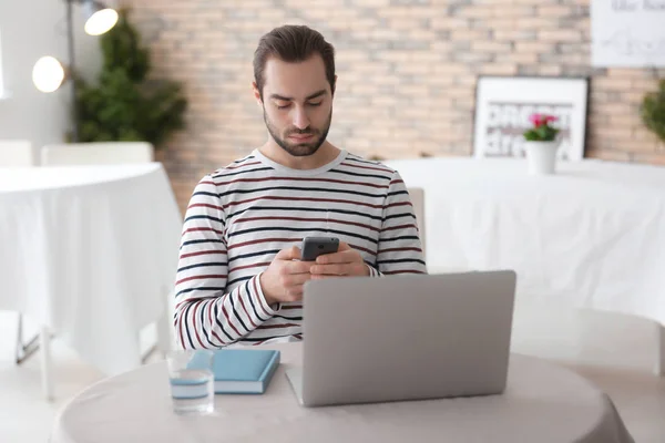 Male freelancer using smartphone while working with laptop in home office — Stock Photo, Image