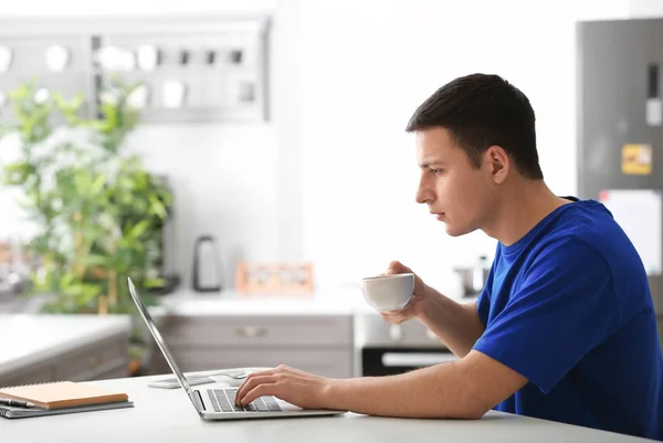 Hombre joven bebiendo café mientras trabaja con el ordenador portátil en la oficina en casa — Foto de Stock