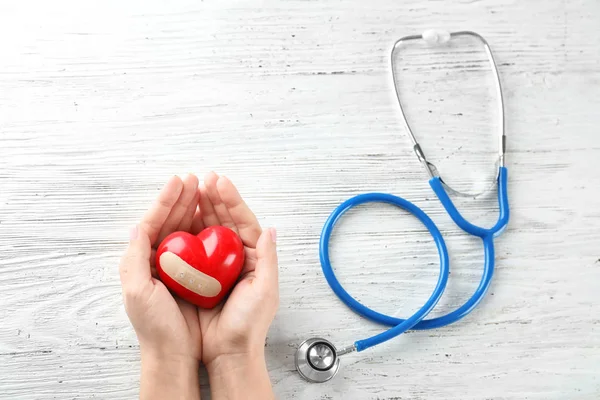 Woman holding red heart with medical plaster on wooden background. Health care concept — Stock Photo, Image