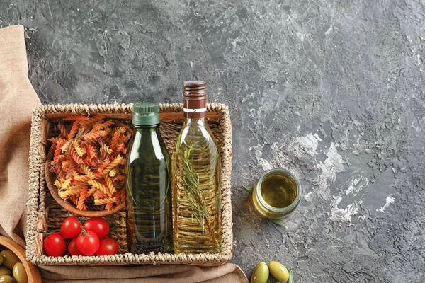 Rieten mand met flessen van olijfolie, pasta en tomaten op grijze achtergrond — Stockfoto