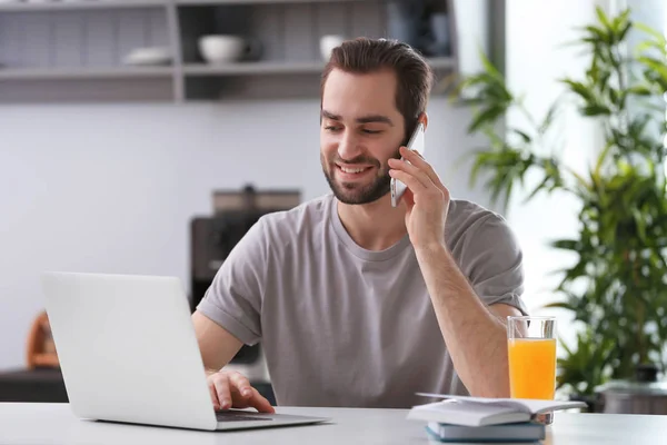 Αρσενικό freelancer μιλάμε στο τηλέφωνο ενώ εργάζεστε με το φορητό υπολογιστή στο γραφείο στο σπίτι — Φωτογραφία Αρχείου