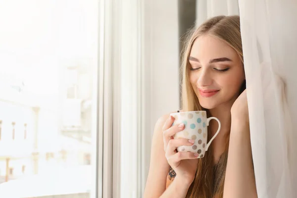 Mujer joven bebiendo café de pie cerca de la ventana en casa — Foto de Stock