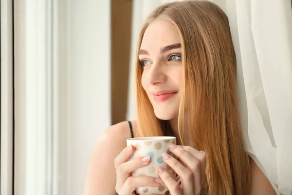 Young woman drinking coffee while standing near window at home — Stock Photo, Image