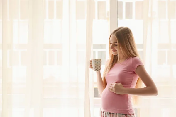 Mladá těhotná žena pití čaje když stál blízko okna doma — Stock fotografie